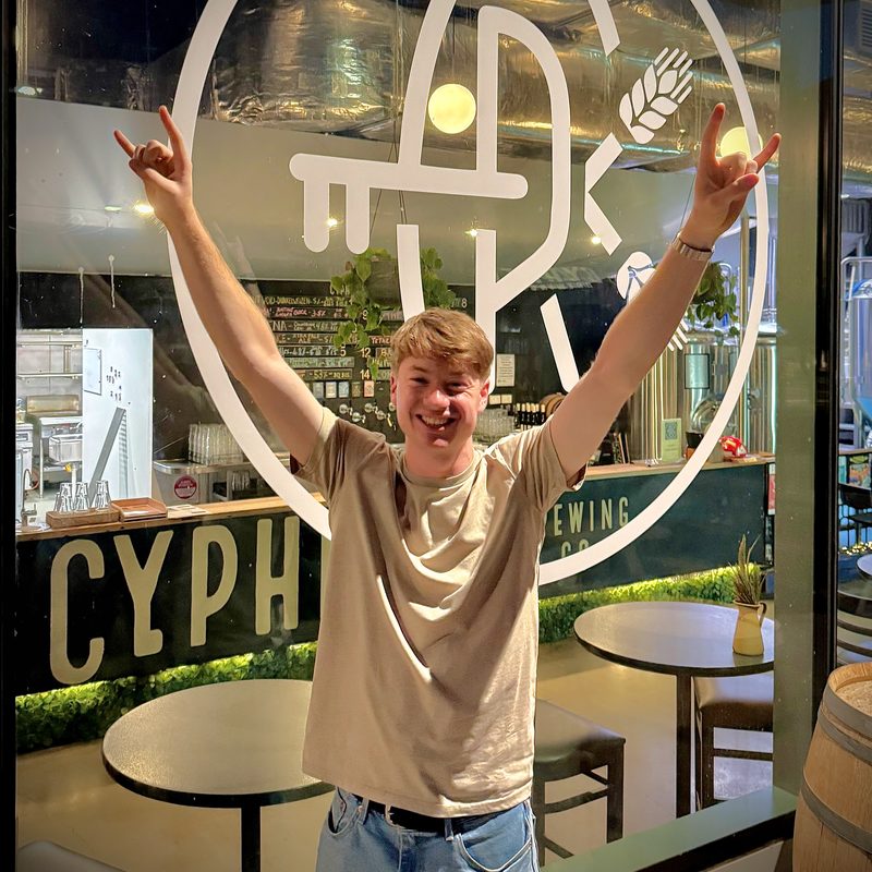 Damien Granet grinning with both arms raised outside Cypher Brewing Co.